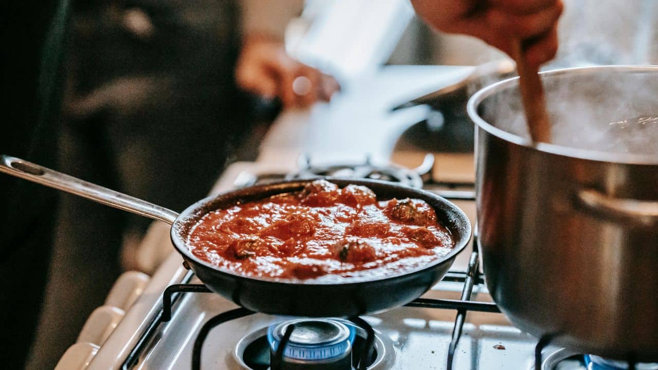 A skillet of meatballs in tomato sauce on a gas stove, next to a pot being stirred.