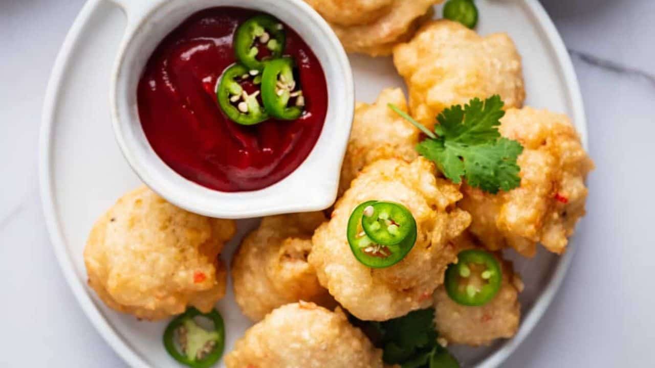 A plate of fried fritters garnished with sliced green chili peppers, accompanied by a small dish of red sauce, and garnished with cilantro.