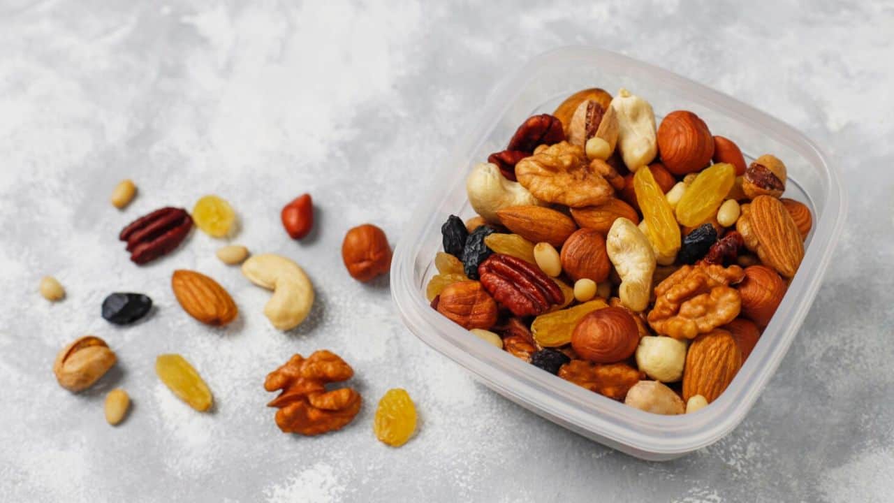 Plastic container filled with mixed nuts and dried fruits, with a few scattered outside on a gray surface.