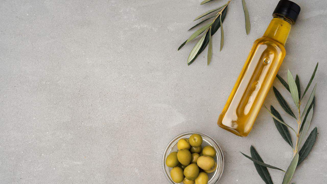 A bottle of olive oil, a small bowl of green olives, and olive branches arranged on a gray surface.