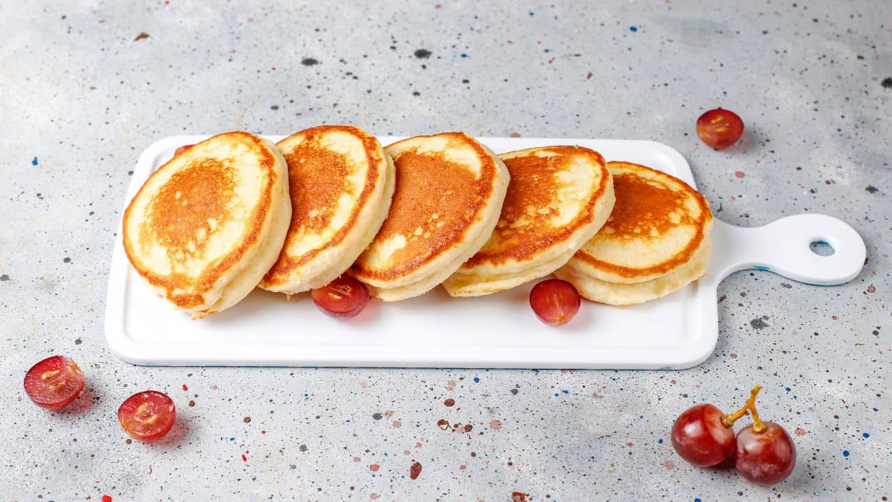 Five fluffy pancakes arranged on a white rectangular board with scattered red grapes on a speckled surface.