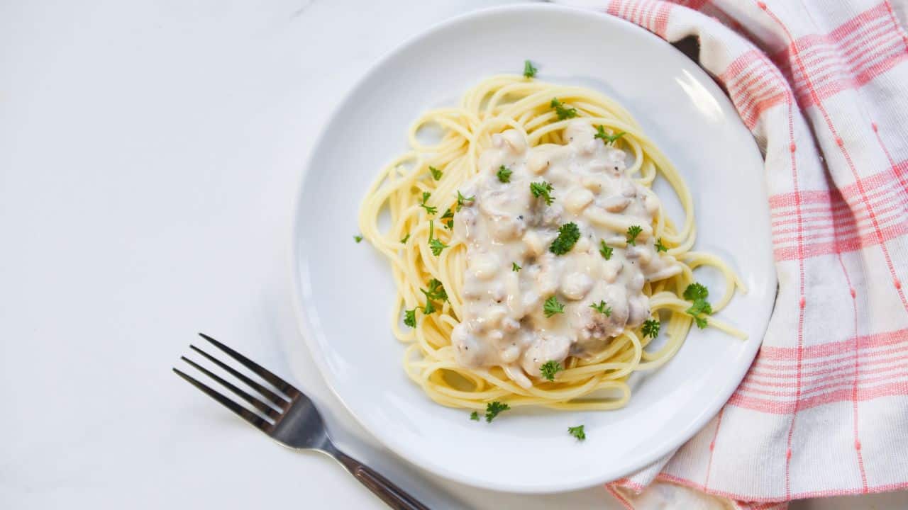 A plate of spaghetti with white sauce and parsley garnish. A fork and red-striped napkin are beside the plate on a white surface.