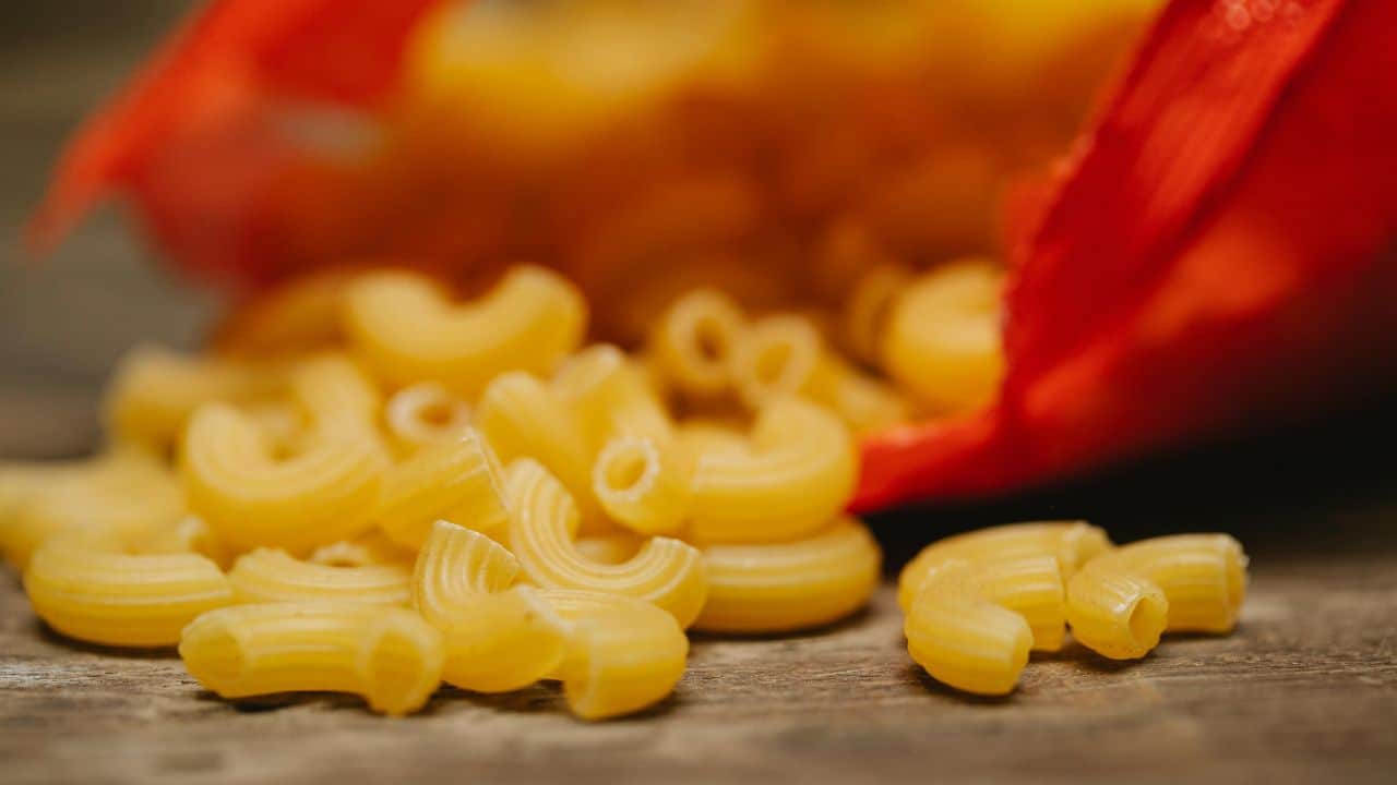 Close-up of uncooked elbow macaroni spilling from a red package onto a wooden surface.