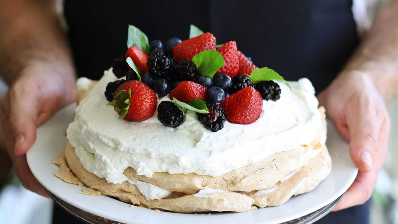 A person holds a pavlova topped with whipped cream, strawberries, blackberries, blueberries, and mint leaves on a white plate.