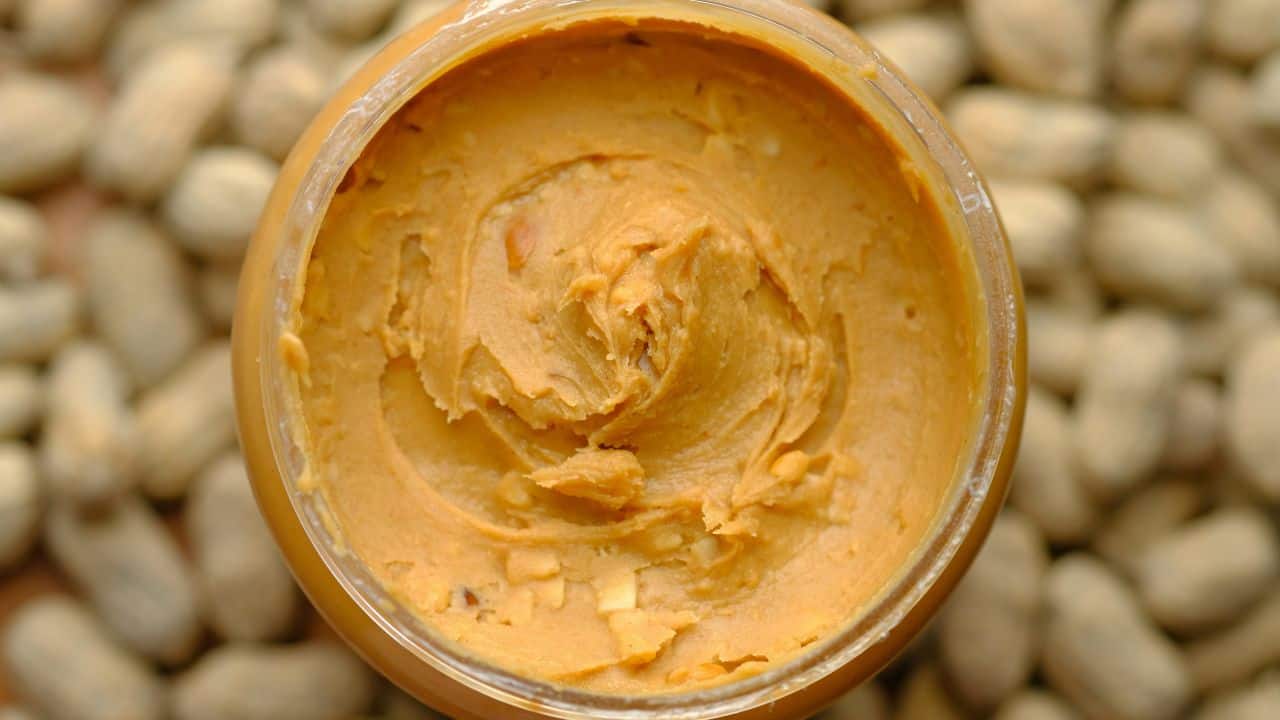 An open jar of peanut butter with a smooth texture, surrounded by unshelled peanuts.