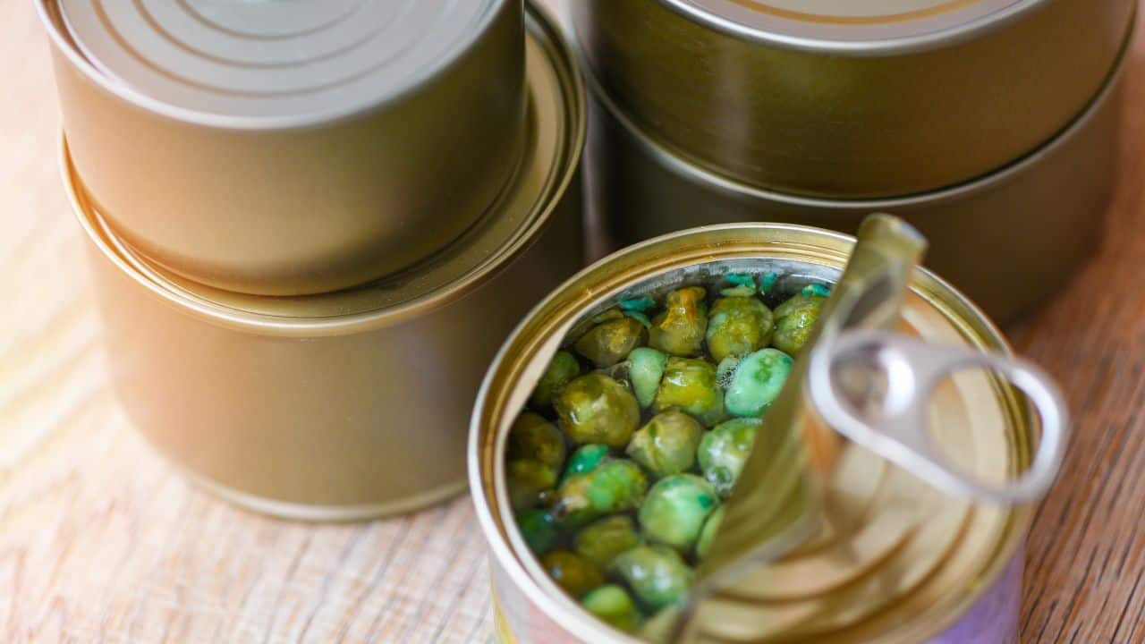 A stack of closed tin cans sits beside an open can containing spoiled green peas with mold, on a wooden surface.