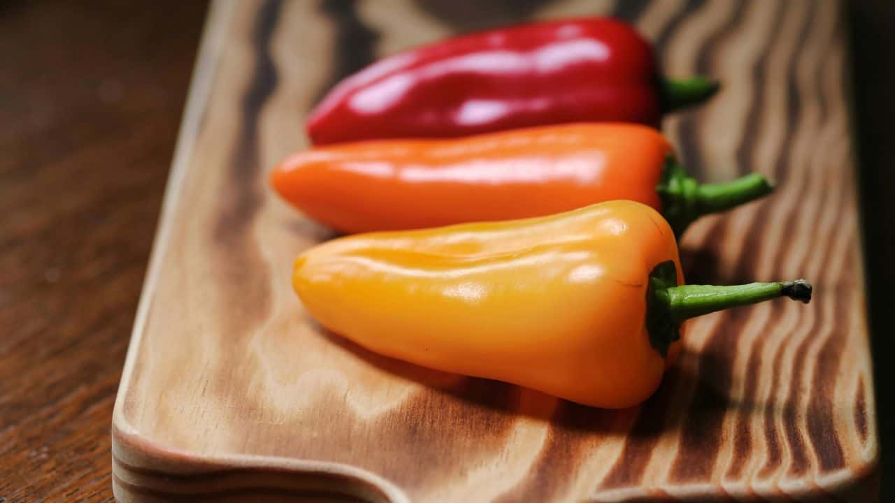 Three mini bell peppers in red, orange, and yellow are placed on a wooden cutting board.