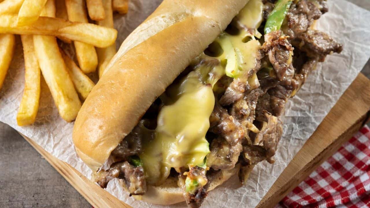 A cheesesteak sandwich with melted cheese and meat filling, served on a wooden board with a side of French fries.