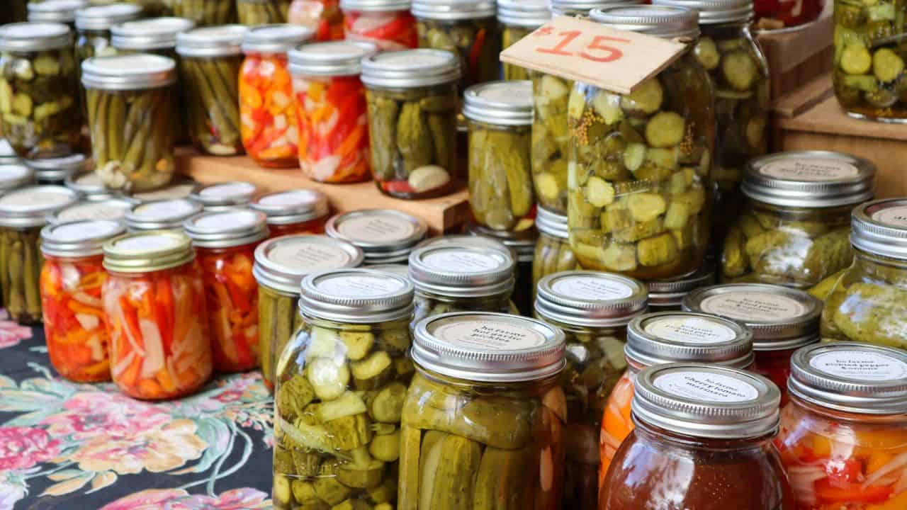 Jars of assorted pickled vegetables such as cucumbers and peppers are displayed on a table at a market. A sign with the number 15 is visible.