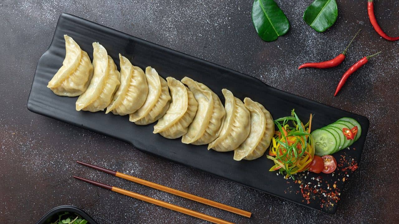 A row of dumplings on a black rectangular plate, garnished with sliced cucumber, chili, and vegetables. Red chili peppers and green leaves are scattered around the surface. Chopsticks are placed nearby.