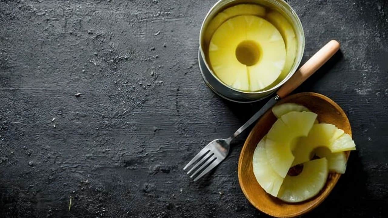 Open can of pineapple slices with a fork beside it and more slices on a wooden plate, placed on a dark textured surface.