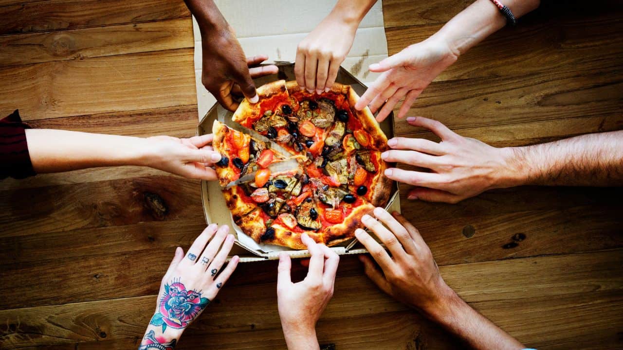 Several hands reaching for slices of pizza in a box on a wooden table.
