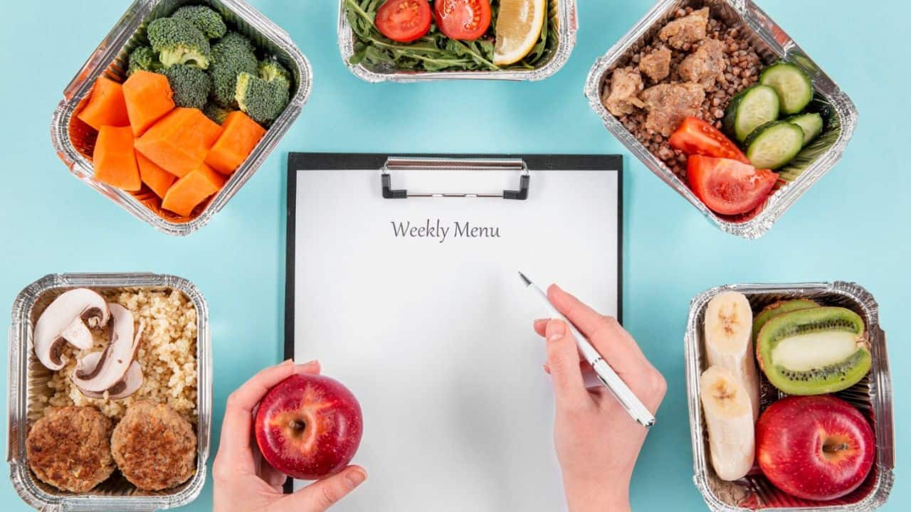 A person writes a weekly menu on a clipboard surrounded by meal prep containers with vegetables, grains, and fruits.