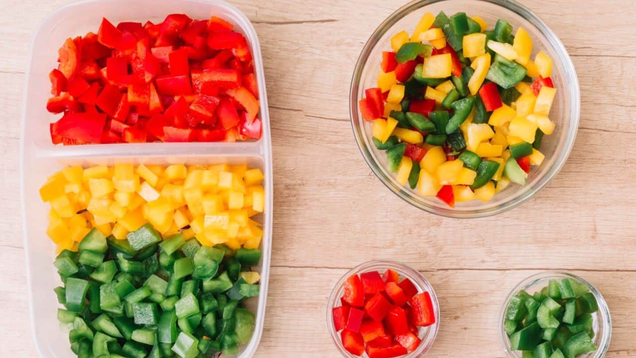 Chopped red, yellow, and green bell peppers in bowls and a container on a wooden surface.