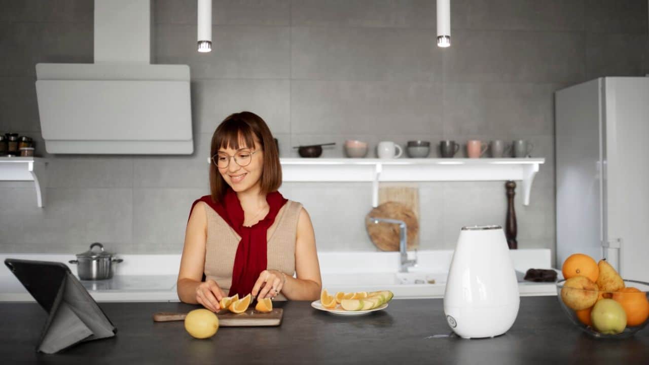 Woman in a kitchen slicing citrus fruits while looking at a tablet, with a fruit bowl and a kitchen appliance on the counter.