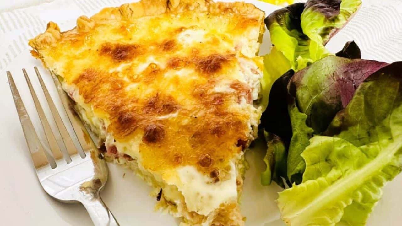 Slice of Quiche Lorraine on white plate next to fork and green salad.