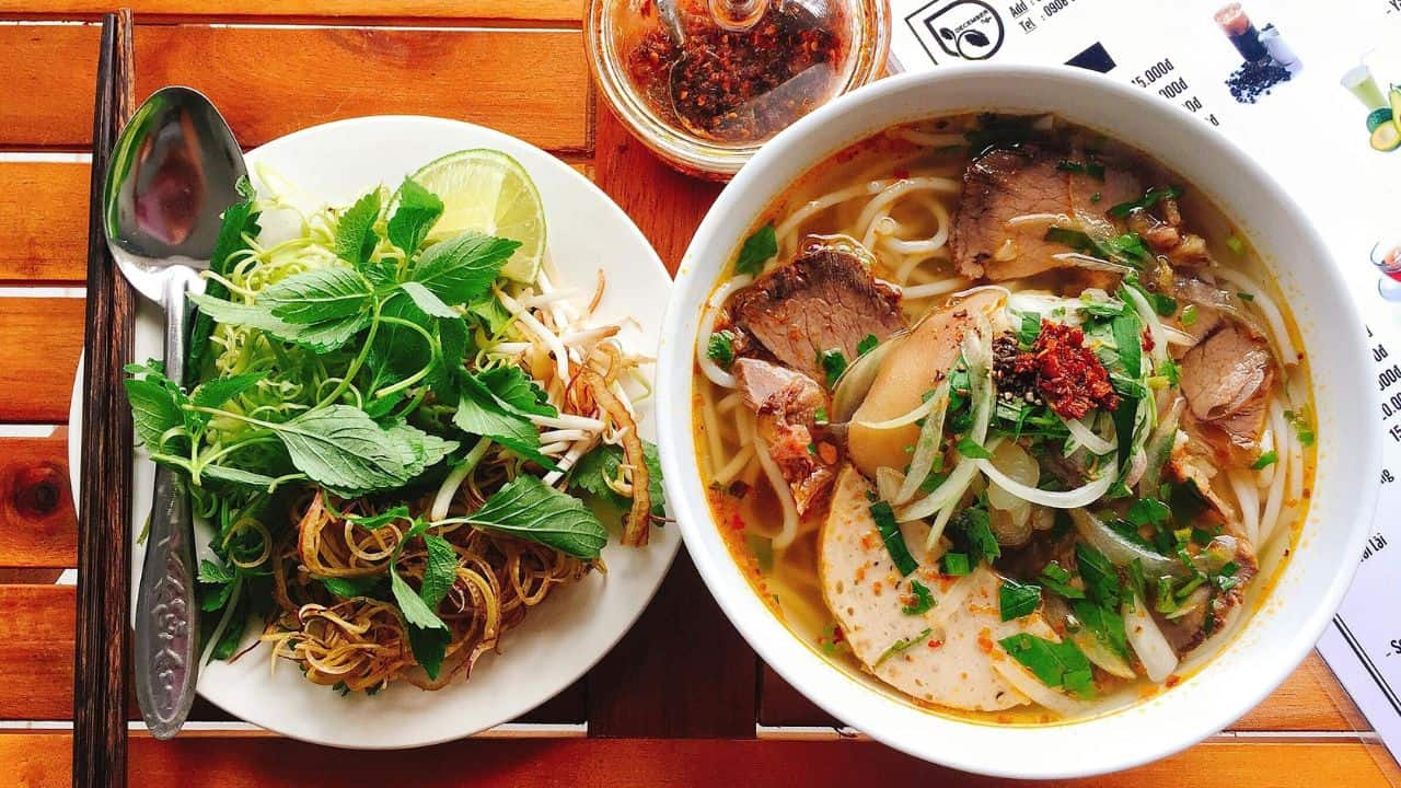 A bowl of noodle soup with meat and vegetables next to a plate of herbs, lime, and bean sprouts on a wooden table.