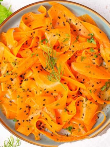 A plate of carrot ribbons sprinkled with black sesame seeds, garnished with dill, and accompanied by a fork and knife with yellow handles on a light surface.