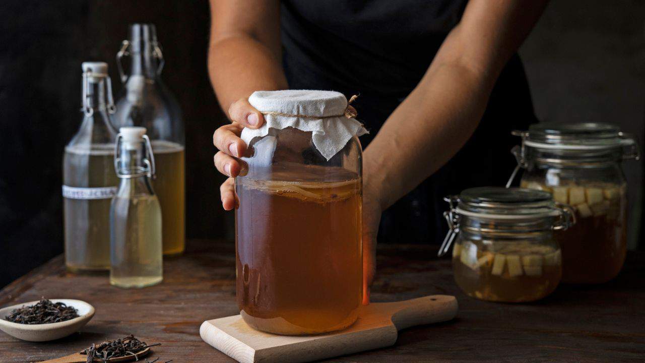 A person handling a large glass jar of fermenting liquid on a wooden table, surrounded by bottles and jars of ingredients.