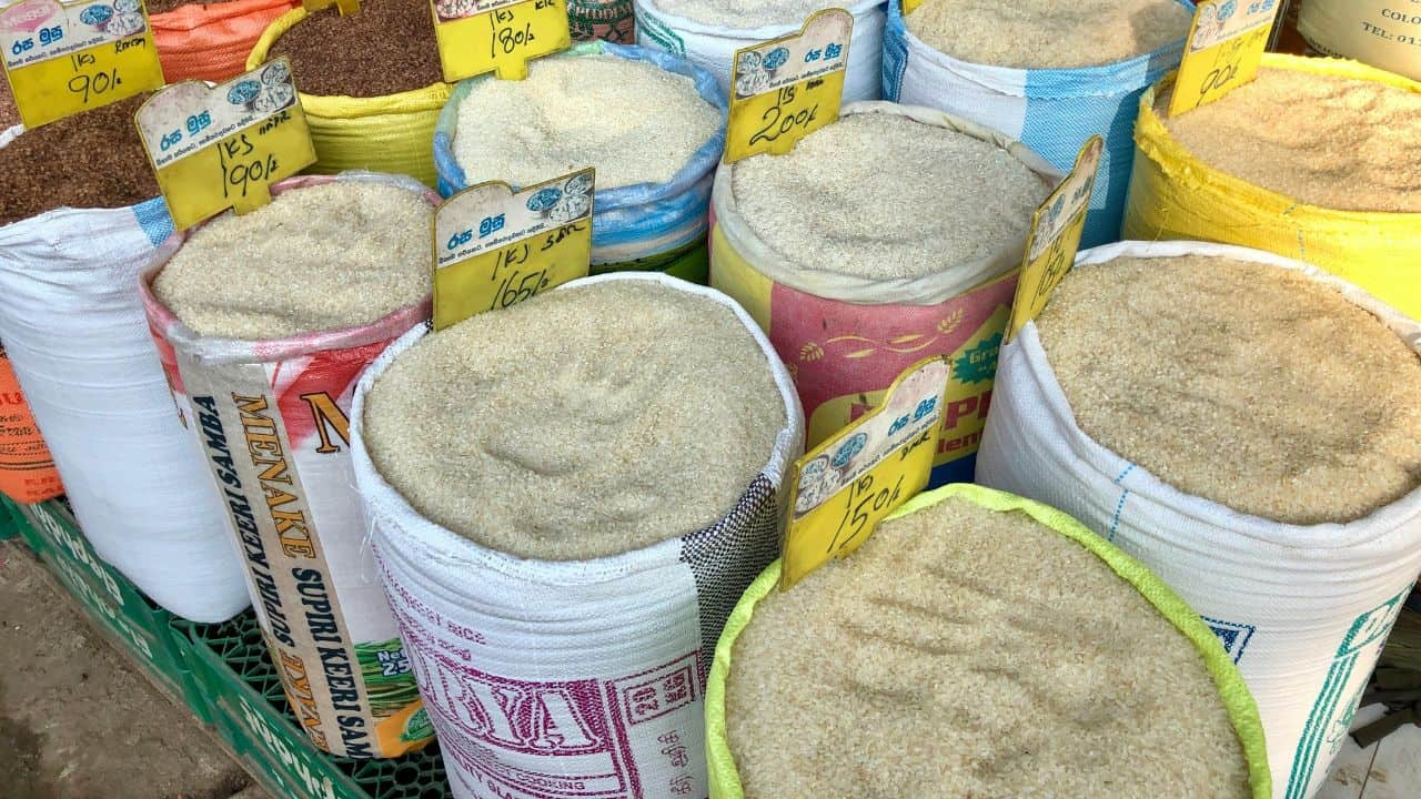 Bags filled with various grains and powders are displayed for sale, each labeled with a price and name in a market setting.
