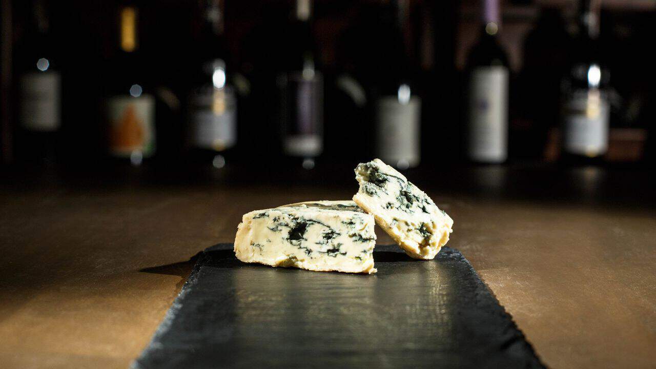 Two pieces of blue cheese on a dark slate board with blurred wine bottles in the background.