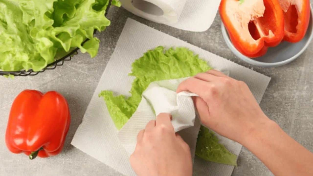 Hands drying lettuce leaves with a paper towel next to red bell peppers on a countertop.