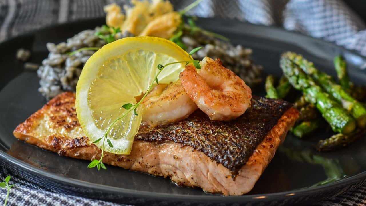 Grilled salmon topped with shrimp and lemon slices, served with asparagus and a side of black pasta.