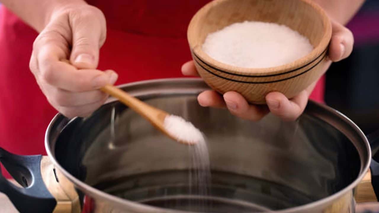 Person adding salt to a pot of water using a wooden spoon, while holding a small wooden bowl filled with salt.