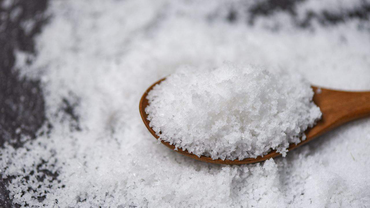 A wooden spoon holding coarse salt is placed on a dark surface sprinkled with more salt.