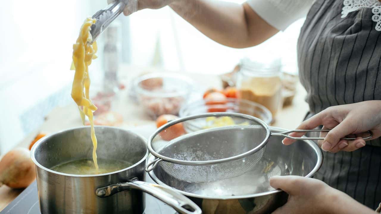 Two people preparing pasta: one uses tongs to lift noodles from a pot, while the other holds a strainer over a bowl in a kitchen setting with various ingredients nearby.