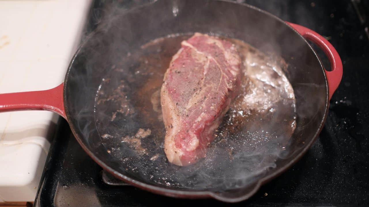 A steak is sizzling in a red cast iron skillet on a stovetop, creating smoke.