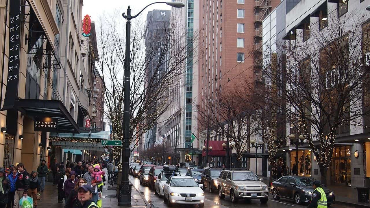 Busy city street with cars, pedestrians, and buildings lining both sides. Trees are decorated with string lights, and people are walking on the sidewalks. Cloudy sky above.