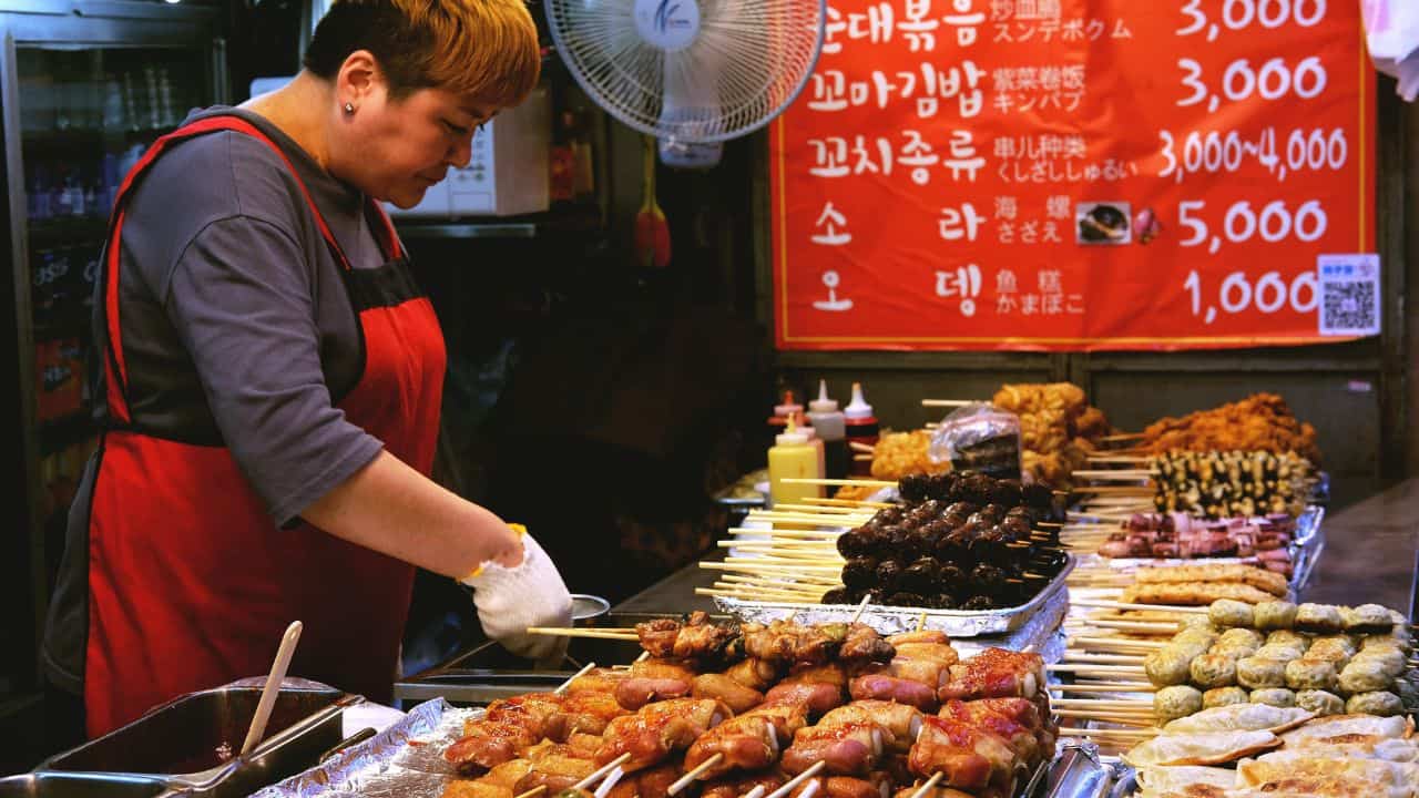 Vendor in a red apron selling skewered meats and snacks at a street food stall with a menu in Korean displayed in the background.