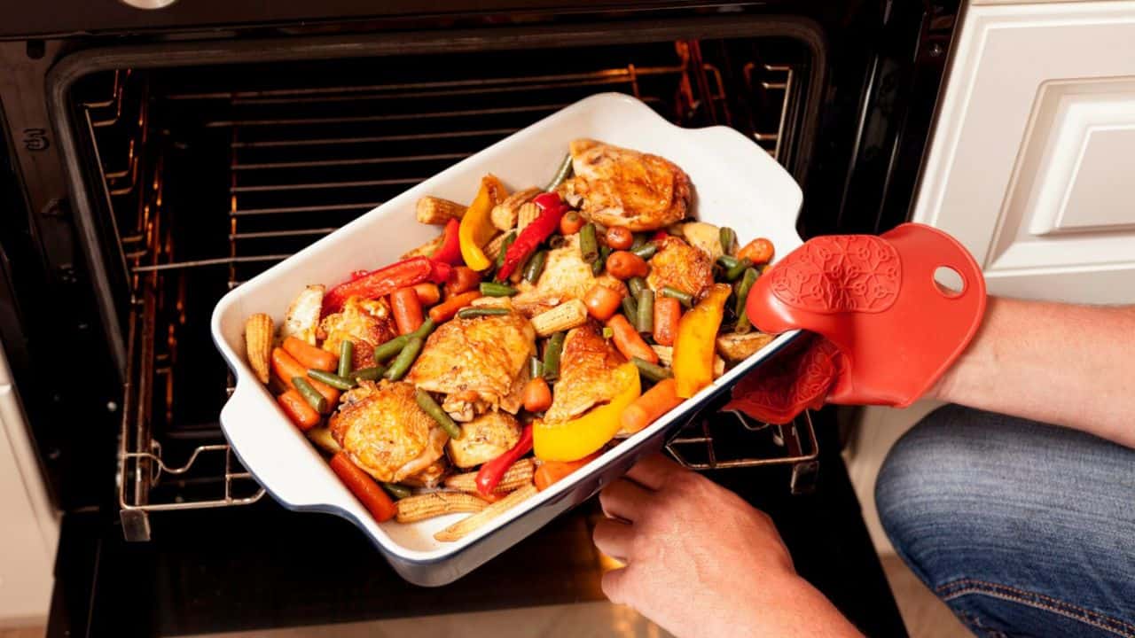 Person removing a baking dish with roasted chicken and mixed vegetables from an oven using a red oven mitt.