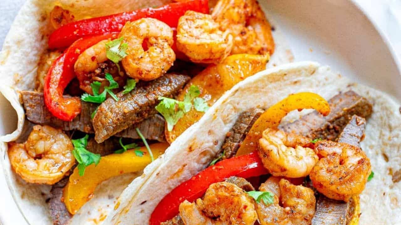 Two tacos filled with shrimp, steak strips, red and yellow bell peppers, garnished with cilantro on a white plate.