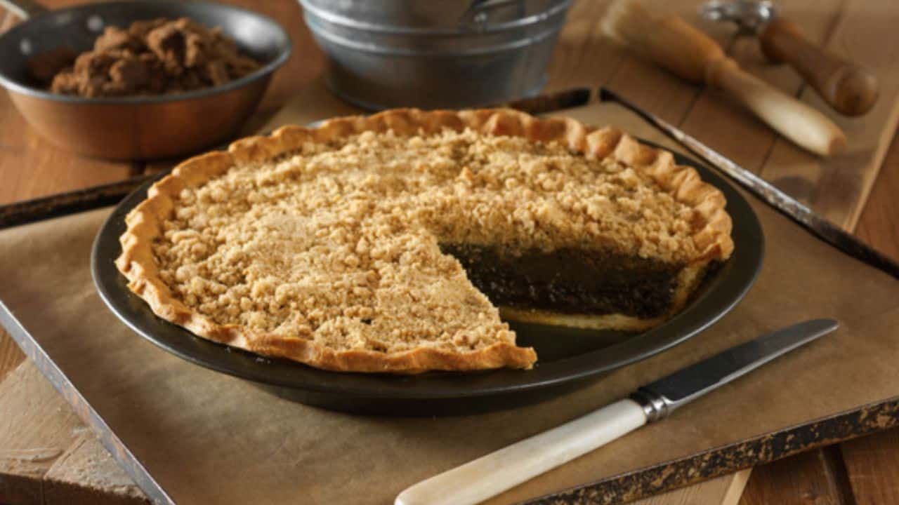 A pie with a golden brown crumb topping sits on a baking tray. A slice has been removed, revealing a dark filling. A knife is placed next to the pie.
