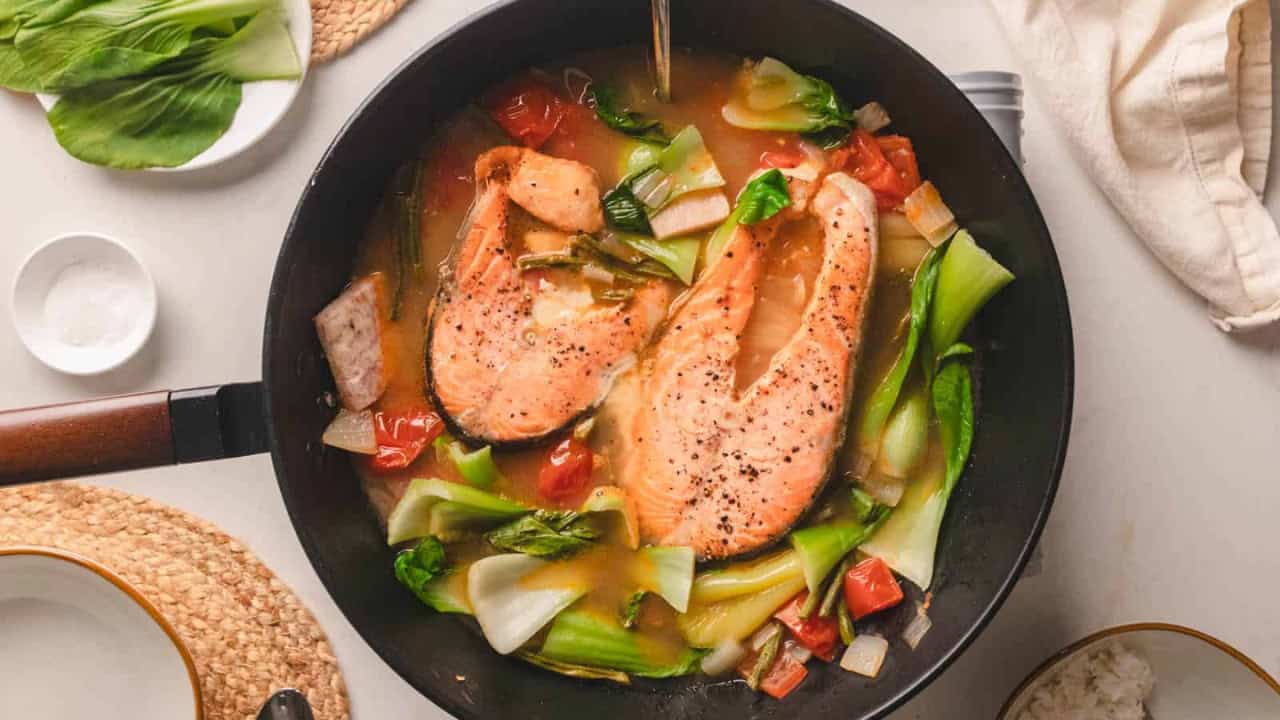 Two salmon steaks cooking in a pan with bok choy, tomatoes, and onions in a broth.