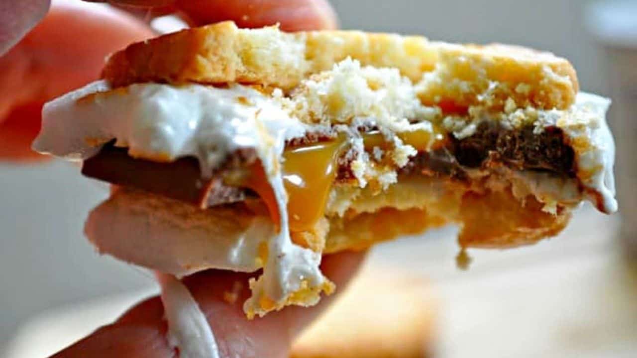 Close-up of a person holding a graham cracker sandwich with marshmallow, chocolate, and caramel filling.