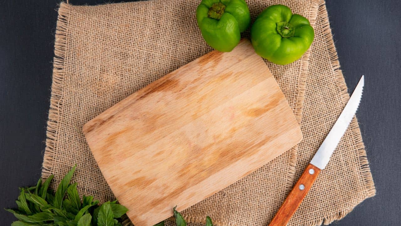 Wooden cutting board on burlap with two green bell peppers, a serrated knife, and fresh herbs on a dark surface.