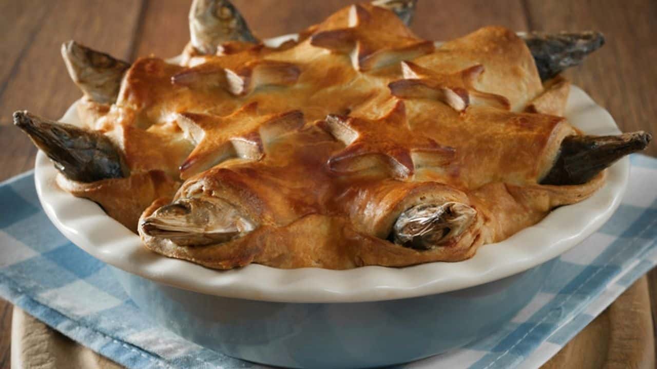 A fish pie with whole fish heads protruding from a golden, star-shaped pastry crust sits in a white dish on a blue and white checkered cloth.