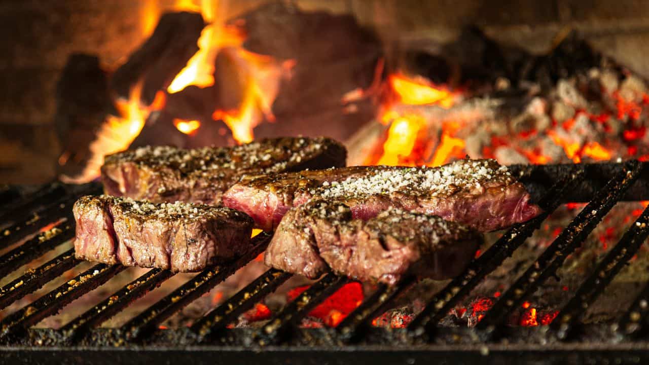 Steaks sizzling on a grill over hot coals, with flames in the background.