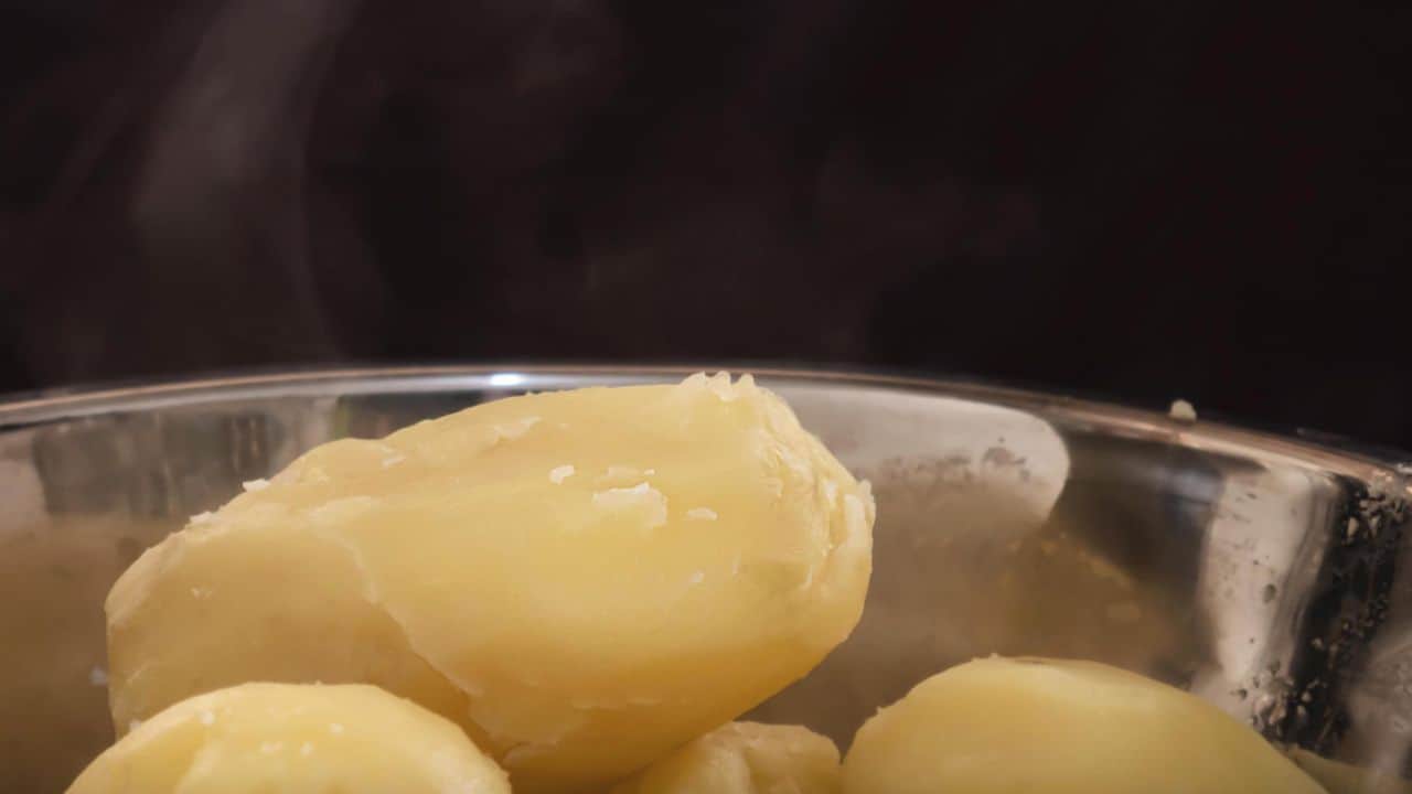 Close-up of boiled potatoes in a stainless steel pot with steam rising against a dark background.