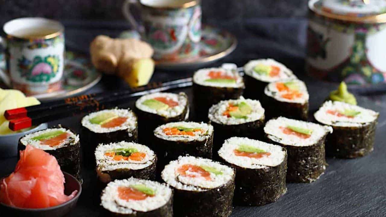 Sushi rolls with salmon, avocado, and cucumber on a slate board, accompanied by a bowl of pickled ginger, chopsticks, and decorative tea cups in the background.