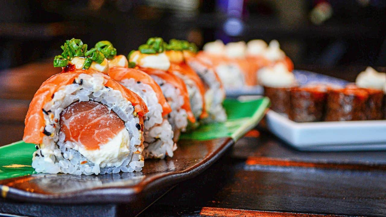 A close-up of sushi rolls topped with salmon, green onions, and sauce on a ceramic tray, with more sushi visible in the background.