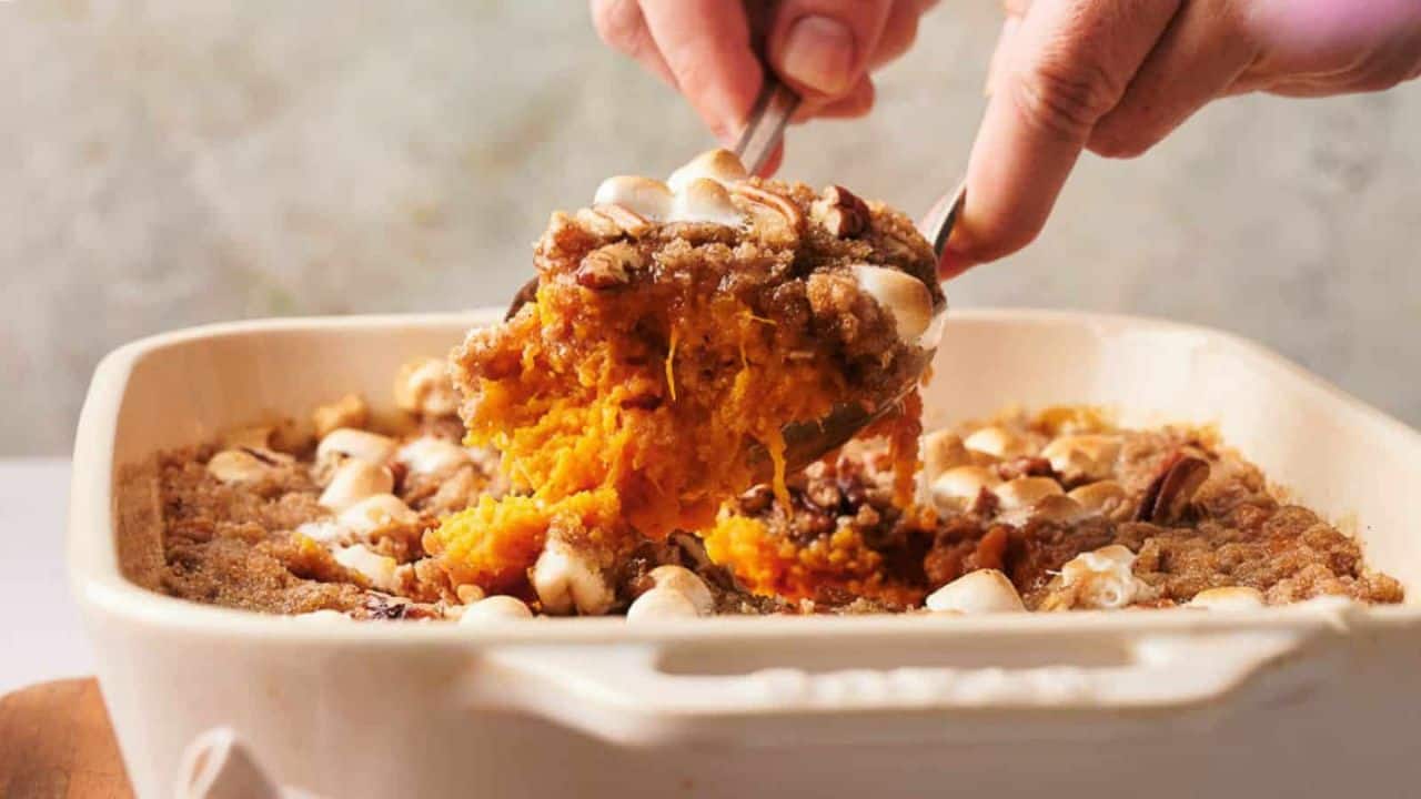 A person scoops a serving of sweet potato casserole topped with marshmallows and pecans from a square baking dish.