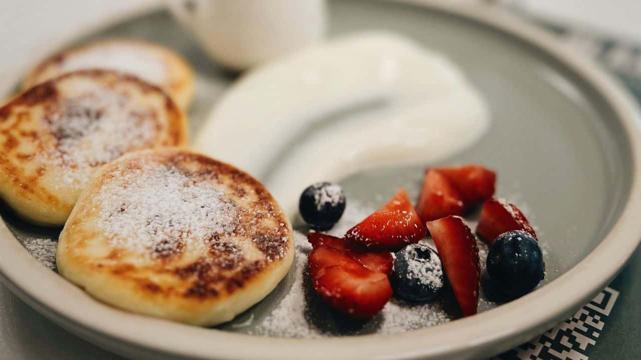 A plate with pancakes dusted with powdered sugar, a dollop of cream, and sliced strawberries and blueberries.