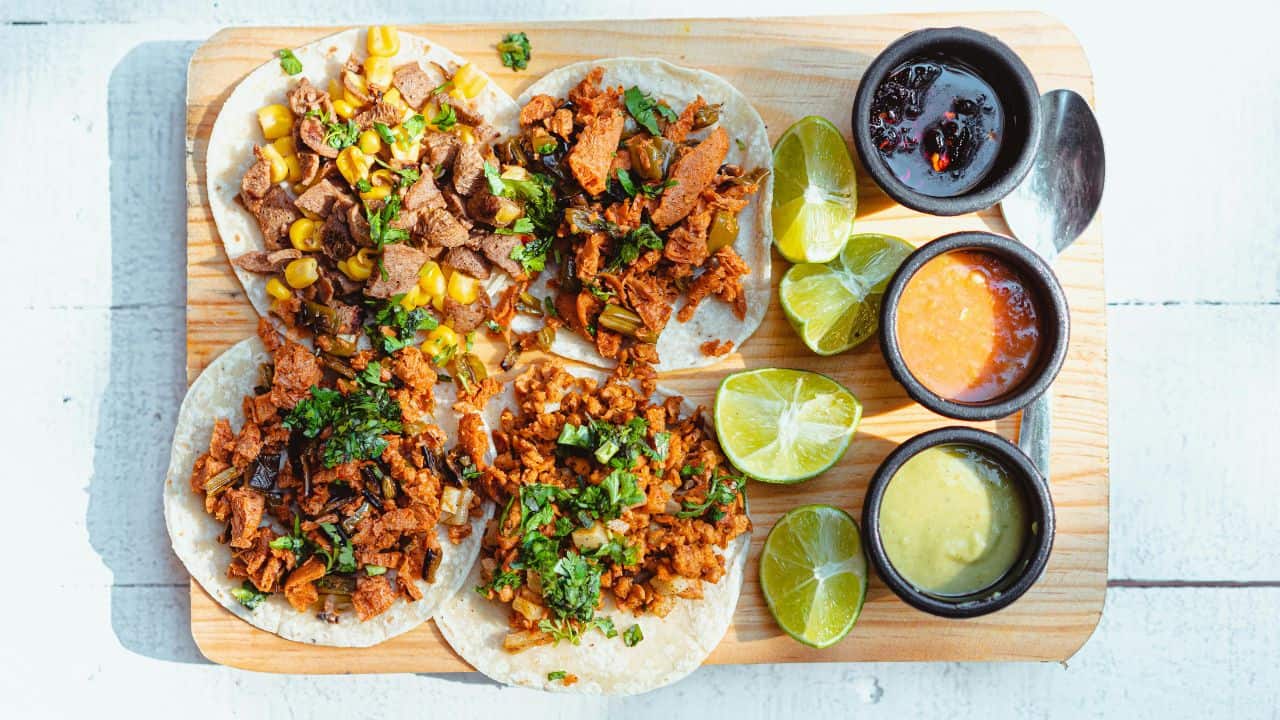 Four tacos with various toppings on a wooden board, accompanied by lime wedges and three small bowls of different salsas.
