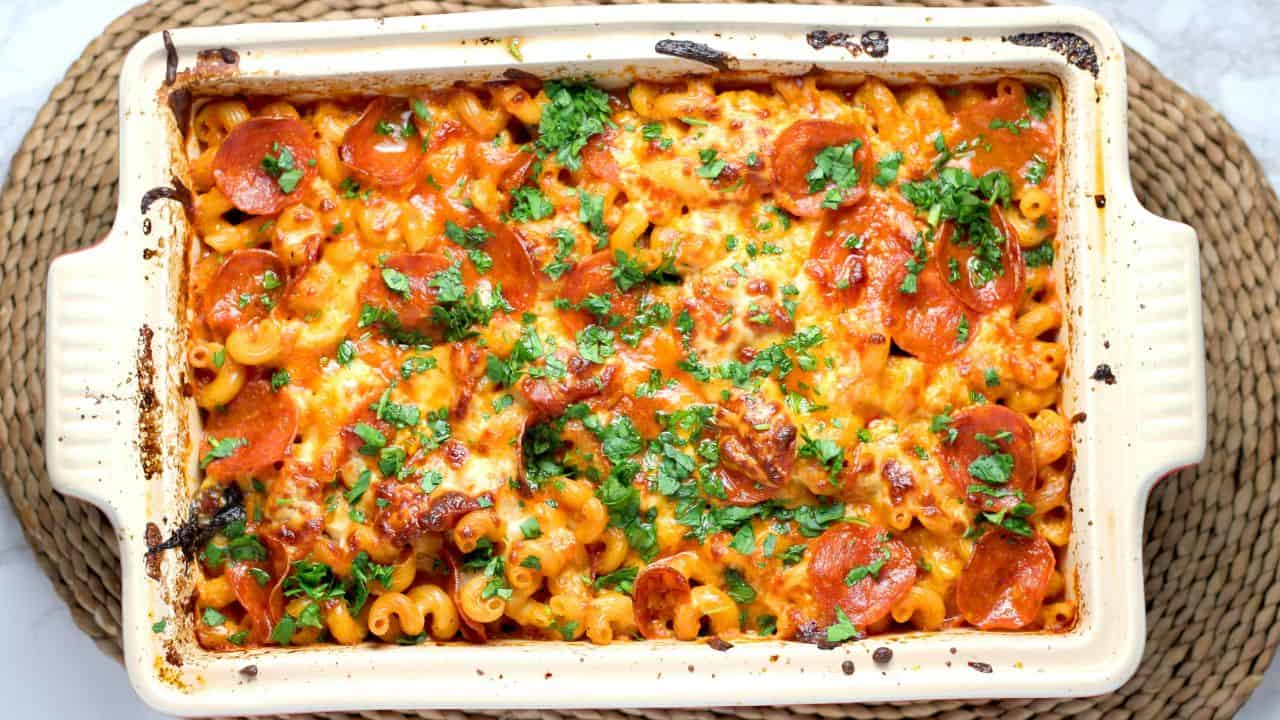 Baked pasta casserole with melted cheese, pepperoni slices, and chopped parsley in a rectangular dish on a woven mat.