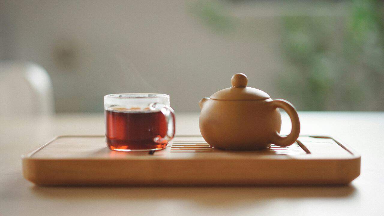 A glass cup of tea and a clay teapot sit on a wooden tray.