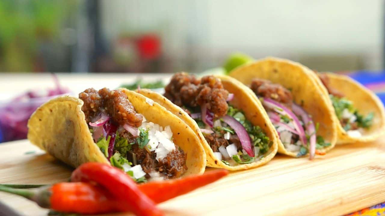 Four tacos filled with meat, onions, and cilantro are lined up on a wooden board with red chili peppers in the foreground.